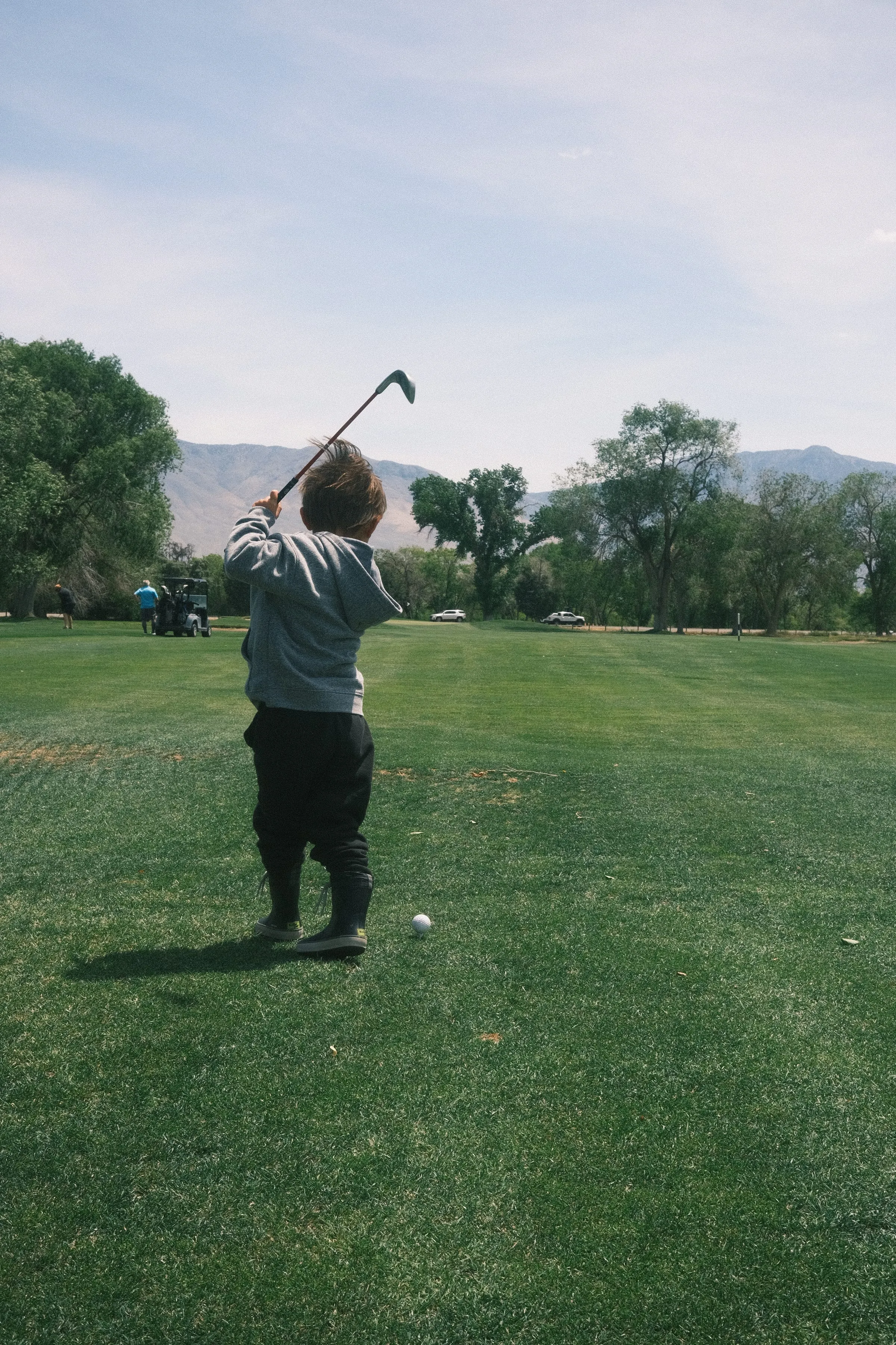 A kid mid-backswing on a golf fairway, ball waiting at his feet, mountains behind