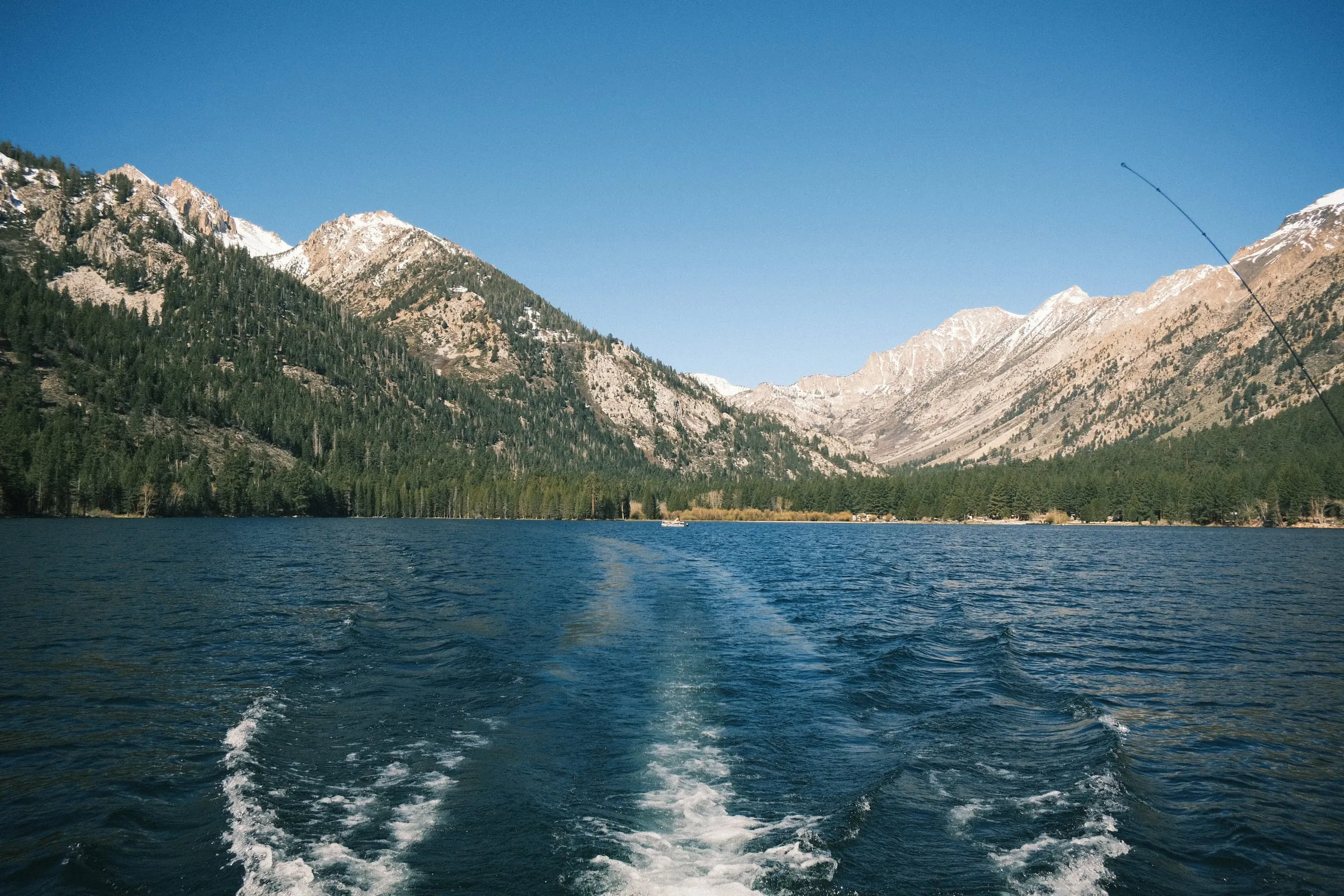Boat wake trailing across a mountain lake, a fishing rod tip in frame, snow-dusted peaks behind